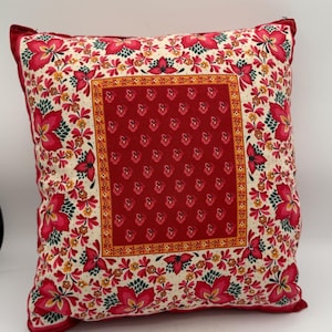 May include: Square decorative pillow with a floral pattern. The pillow features a central red square with a repeating pattern, framed by a yellow border. The outer edges have a cream background with red flowers and green leaves. The pillow has a red border.