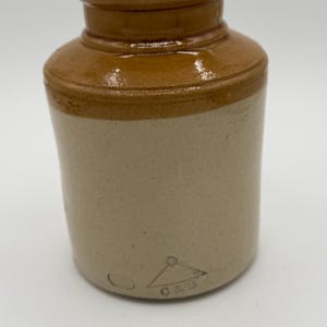 May include: A small, antique stoneware jar with a rounded body and a narrow neck. The jar is primarily beige with a band of brown around the top. A small, triangular marking with the letters "C & B" is visible on the side.