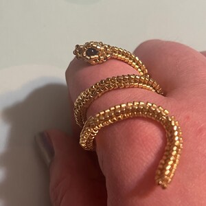 May include: A gold snake ring coiled around a finger. The ring is made of small, connected beads, with a snake head featuring a dark eye. The ring's design wraps around the finger, creating a unique and eye-catching accessory.
