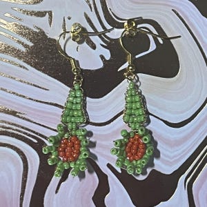 May include: A pair of handmade beaded earrings. Each earring features a gold-colored hook, a green beaded top section, and a red and green beaded floral-like design. The earrings are displayed against a marbled background with gold accents.