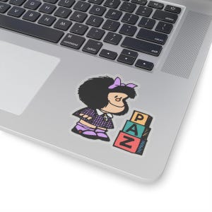 May include: A sticker featuring a cartoon girl with black hair and a purple dress standing next to a stack of colorful blocks that spell out "PAZ" in Spanish.