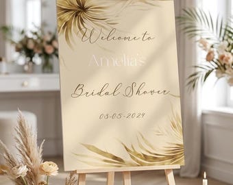 May include: A rectangular cream-colored sign displayed on a wooden easel. The sign features gold-toned palm leaf illustrations and black script text that reads Welcome to Amelia's Bridal Shower 03-05-2024. The design is clean and centered.