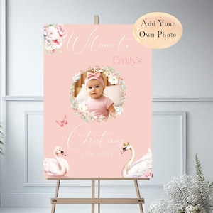 May include: A pink welcome sign for a christening, featuring a baby's photo surrounded by a floral wreath. The sign includes the text "Welcome to Emily's Christening 2 March 2024" and illustrations of swans and a butterfly.
