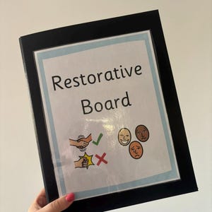 May include: A black binder featuring a white insert titled Restorative Board. The insert displays a light blue border, a graphic of two hands shaking with a green checkmark, a fist bump with a red X, and three circular facial expressions.