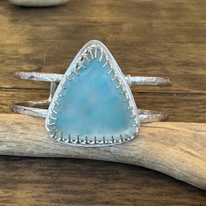 May include: A silver cuff bracelet with a triangular, light blue stone. The stone is framed with silver and has a textured surface. The bracelet is resting on a piece of driftwood.