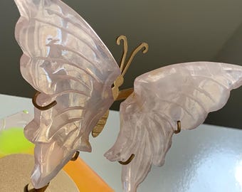 May include: A decorative butterfly sculpture with carved wings. The wings are a translucent pink, and the body and antennae are gold-colored. The butterfly is mounted on a gold-colored base, with a colorful background.