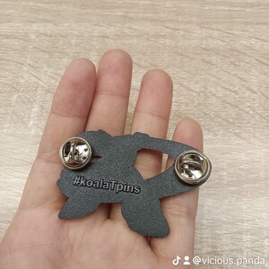 Op de afbeelding: Zwarte emaille pin met een koala die een hart vasthoudt met de tekst "#koalaTpins" op de achterkant. De pin heeft een zilveren metalen sluiting.