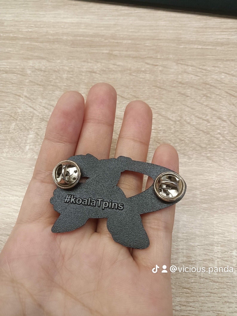 Op de afbeelding: Zwarte emaille pin met een koala die een hart vasthoudt met de tekst "#koalaTpins" op de achterkant. De pin heeft een zilveren metalen sluiting.