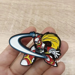 Op de afbeelding: Een emaille pin met een personage uit de Mega Man X-videogameserie. Het personage wordt afgebeeld in een dynamische pose, zwaaiend met een groot, sikkelvormig energiemes. De pin heeft een geel en wit kleurenschema, met een blauwe omtrek rond het mes.