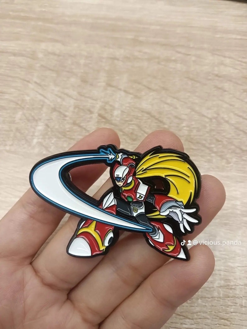 Op de afbeelding: Een emaille pin met een personage uit de Mega Man X-videogameserie. Het personage wordt afgebeeld in een dynamische pose, zwaaiend met een groot, sikkelvormig energiemes. De pin heeft een geel en wit kleurenschema, met een blauwe omtrek rond het mes.