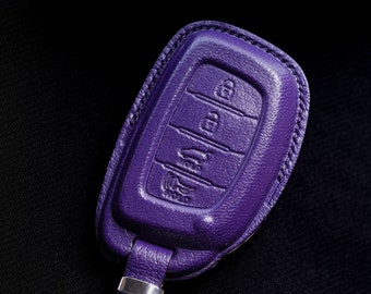 May include: A purple leather car key fob cover with visible stitching. The cover features embossed icons for locking, unlocking, trunk release, and a panic alarm button. A matching purple leather strap with a silver metal snap and ring is attached.