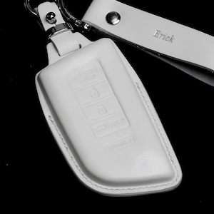 May include: A white leather key fob cover with a matching wrist strap and silver-tone hardware. The key fob cover has embossed buttons and a stitched border. The wrist strap is engraved with the name "Erick".