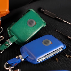 May include: Leather key fob covers in orange, green, and blue, each with a silver-tone accent and a key ring. The key fobs have a rectangular shape with rounded edges. Small tools and hardware are scattered around the key fobs.
