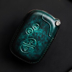 May include: A teal leather key fob cover with embossed key symbols. The cover has a distressed finish and is surrounded by small tools and hardware on a dark surface. The key fob cover is rectangular with rounded edges.