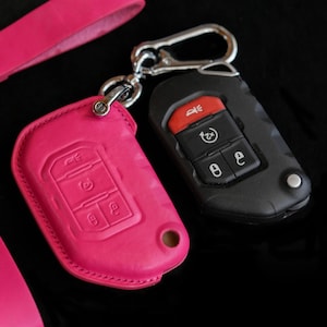 Peut inclure: Un étui de clé en cuir rose vif et un porte-clés noir, reliés par un mousqueton argenté. Le couvercle rose présente des symboles de clé en relief. Le porte-clés noir a des boutons rouges et noirs. Les articles sont sur une surface noire.