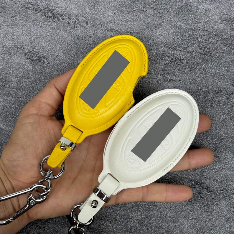 May include: Two leather key fob covers, one yellow and one white, with silver-tone hardware. The covers are oval-shaped with a strap and a metal clasp. The key fobs are shown against a gray background.