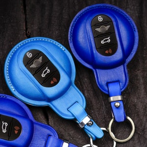 Può includere: Tre custodie blu in pelle per portachiavi per auto. Le custodie sono in tonalità di blu, con un portachiavi e un cinturino. I portachiavi hanno pulsanti neri con icone bianche. Le cuciture sono bianche.