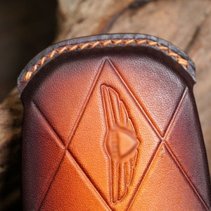 May include: Close-up of a brown leather case with a diamond pattern and a wing emblem. The case has a gradient color from dark brown to light brown. The edges are stitched with orange thread.