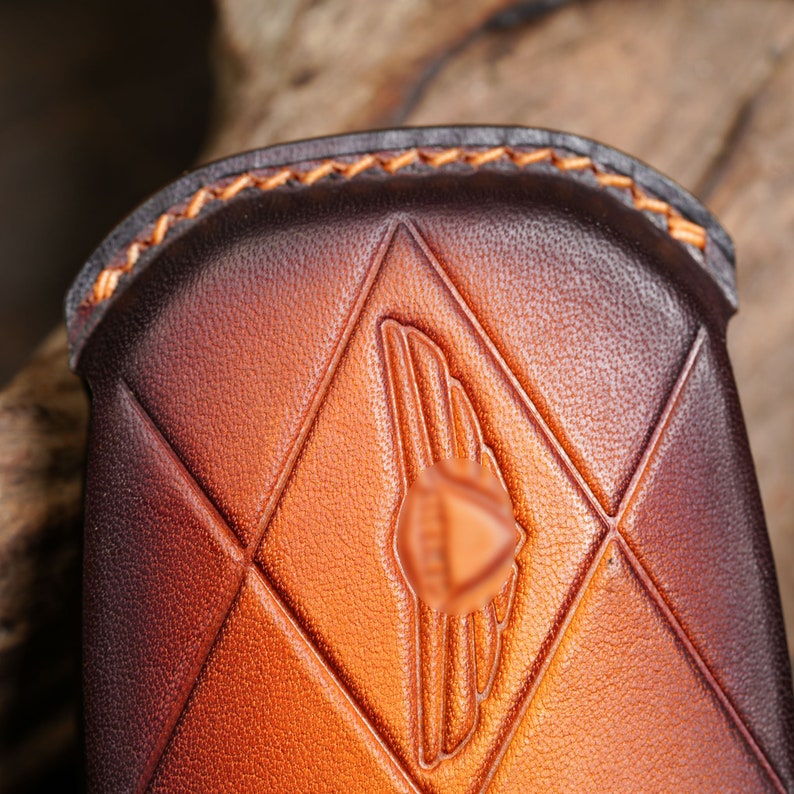 May include: Close-up of a brown leather case with a diamond pattern and a wing emblem. The case has a gradient color from dark brown to light brown. The edges are stitched with orange thread.