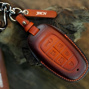 May include: A brown leather car key fob cover with orange stitching and a silver metal clip. The cover features embossed button icons and a small leather tag with the white text JHCN attached to the silver ring hardware.