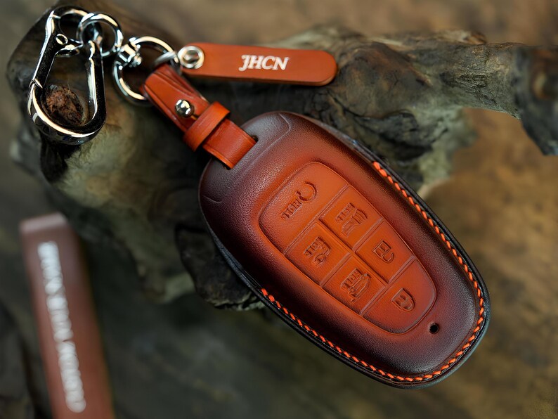 May include: A brown leather car key fob cover with orange stitching and a silver metal clip. The cover features embossed button icons and a small leather tag with the white text JHCN attached to the silver ring hardware.