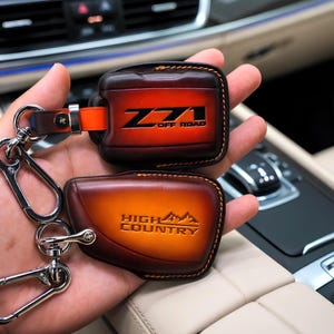 May include: Two leather key fob holders with silver key rings. One is embossed with "Z71 OFF ROAD" in black, the other with "HIGH COUNTRY" and a mountain graphic. The leather has a gradient of brown and orange.