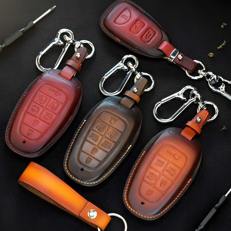 May include: A collection of leather car key fobs in various shades of brown and red. Each fob has a metal clasp and a leather strap. The fobs are arranged on a black surface, with a few tools visible.