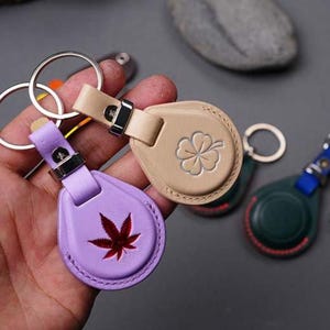 May include: A collection of leather keychains in various colors. One is lavender with a red leaf design, another is tan with a four-leaf clover, and a third is dark green. Each keychain has a metal ring and clasp.