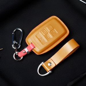 May include: A tan leather key fob case with a red accent, silver carabiner, and matching strap. The case has embossed buttons and a stitched border. The key fob is on a black surface.