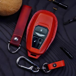 Peut inclure: Un étui de clé de voiture en cuir rouge avec un porte-clés et une dragonne assortis. Le couvercle du porte-clés a des boutons noirs et un emblème argenté. Le porte-clés a un fermoir argenté et une petite sangle en cuir rouge. La dragonne est en cuir rouge.