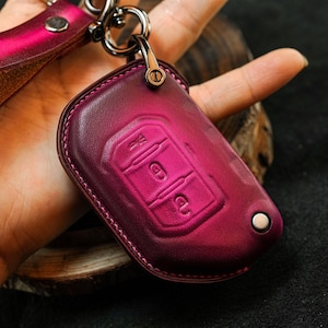 Puede incluir: Una funda para llavero de cuero magenta con un símbolo de llave en relieve en la parte delantera. La funda tiene una correa de cuero a juego y herrajes plateados. La funda para llavero está diseñada para proteger una llave de coche.