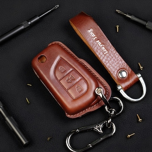May include: A brown leather car key fob cover with a matching key chain and wrist strap. The key fob cover has three buttons and a metal clasp. The wrist strap is embossed with the name "Liuyue Yang". Small screws and screwdrivers are scattered around the key fob.