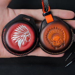 May include: Two leather keyrings with Native American designs. One has a red background with a white profile, the other a brown background with a skull. Both have black and red stitching.