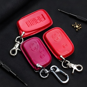 Puede incluir: Tres fundas de cuero para llaveros en rojo y magenta. Cada funda tiene un cierre y un llavero de color plateado. Los llaveros tienen botones en relieve. Un destornillador y pequeños tornillos dorados están en el fondo.