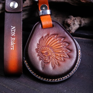 May include: A brown leather keychain with a skull wearing a Native American headdress design. The keychain has a matching leather strap with the text "NDN Riderz" embossed on it. The keychain is attached to a metal ring.