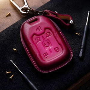 Peut inclure: Un étui de clé de voiture en cuir magenta avec des icônes de boutons en relief. La housse est cousue sur les bords et possède une lanière en cuir et un porte-clés en métal. De petites vis dorées et un tournevis sont éparpillés autour du porte-clés.