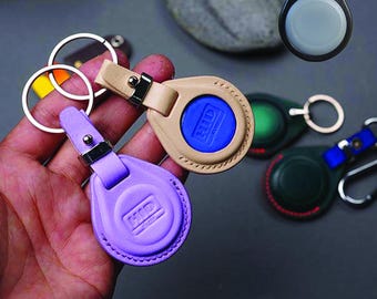 May include: A collection of keychains and AirTag holders in various colors. The holders are made of leather and plastic, with metal key rings and clasps. The colors include purple, tan, black, green, and blue. The AirTag holders have the text "HID" on them.