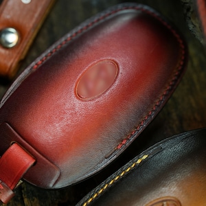 May include: A hand-dyed leather key fob cover featuring a deep red and black gradient finish. The oval-shaped case is stitched with red thread and includes a matching leather strap secured by a silver-toned metal snap button fastener.