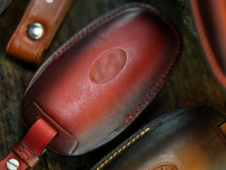 May include: A hand-dyed leather key fob cover featuring a deep red and black gradient finish. The oval-shaped case is stitched with red thread and includes a matching leather strap secured by a silver-toned metal snap button fastener.