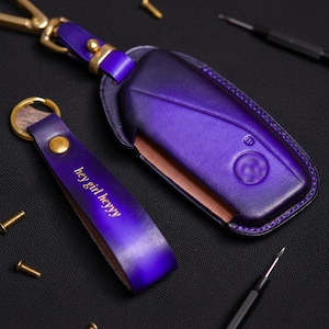 May include: Purple leather car key fob cover and matching keychain. The keychain has the text "hey girl heyyy" in gold lettering. The key fob cover has a gold clasp and a car logo.