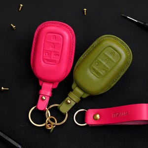 Puede incluir: Dos llaveros de cuero para coche en rosa brillante y verde oliva, con llaveros dorados. El llavero rosa tiene una correa de muñeca a juego con el nombre "Rozalia" en relieve. Los llaveros tienen símbolos de llave en relieve.