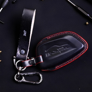 Puede incluir: Funda de llavero de cuero negro con costuras rojas y un llavero a juego. La funda presenta el logotipo de Toyota Supra y las letras "B&P" en la correa. El llavero tiene un cierre plateado.