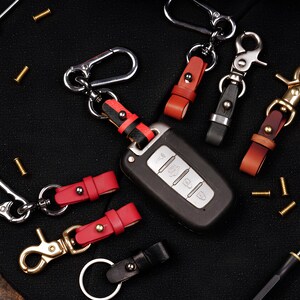 May include: A collection of leather keychains in various colors, including red, black, and brown, with metal clasps and key rings. A car key is also present, showcasing the keychains' functionality. The keychains are designed for securing keys.