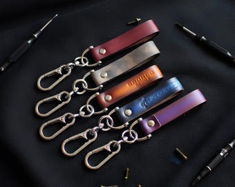 May include: A collection of leather keychains in various colors, including burgundy, camouflage, orange, blue, and purple. Each keychain features a metal clasp and a leather strap with a riveted design. Some keychains have embossed text or a logo.