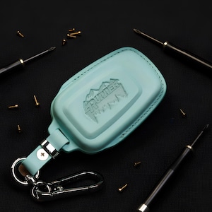 May include: A light blue key fob cover featuring an embossed mountain graphic and the text 4RUNNER. The cover includes a matching light blue strap, a silver-toned metal screw fastener, and a silver-toned metal carabiner clip attached to a ring.
