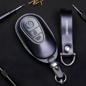 May include: Black leather car key fob cover with embossed buttons and a matching key ring strap. The strap has the number "06763410632" printed on it. Small gold screws and black screwdrivers are in the background.