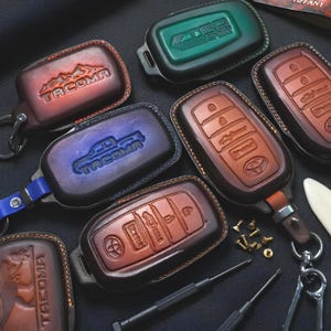 May include: A collection of leather key fob covers in various colors, including brown, green, and blue. Some covers feature embossed designs like mountain ranges, car silhouettes, and the text "TACOMA" and "TRD SPORT".