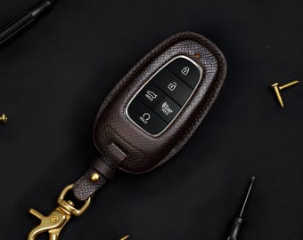 Könnte beinhalten: Eine braune Leder-Autoschlüssel-Hülle mit einem goldfarbenen Verschluss und Schlüsselring. Der Schlüsselanhänger hat schwarze Knöpfe mit weißen Symbolen. Der Hintergrund ist schwarz mit kleinen goldenen Schrauben und zwei schwarzen Schraubendrehern.