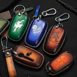 Puede incluir: Una colección de fundas de cuero para llaveros en varios colores, incluyendo verde, azul y marrón. Cada funda presenta un diseño único, como una bandera americana en forma de corazón, una silueta y un diseño de llave de coche. Los llaveros tienen llaveros de metal.