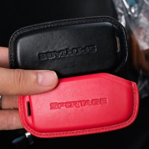 Peut inclure: Deux étuis de clé en cuir, un noir et un rouge, avec le mot "SPORTAGE" en relief. Le noir est au-dessus du rouge. Les étuis sont tenus dans une main. Mesures non disponibles.